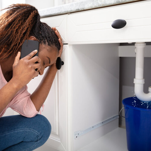 young woman calling an emergency plumber while dealing with a leaking pipe